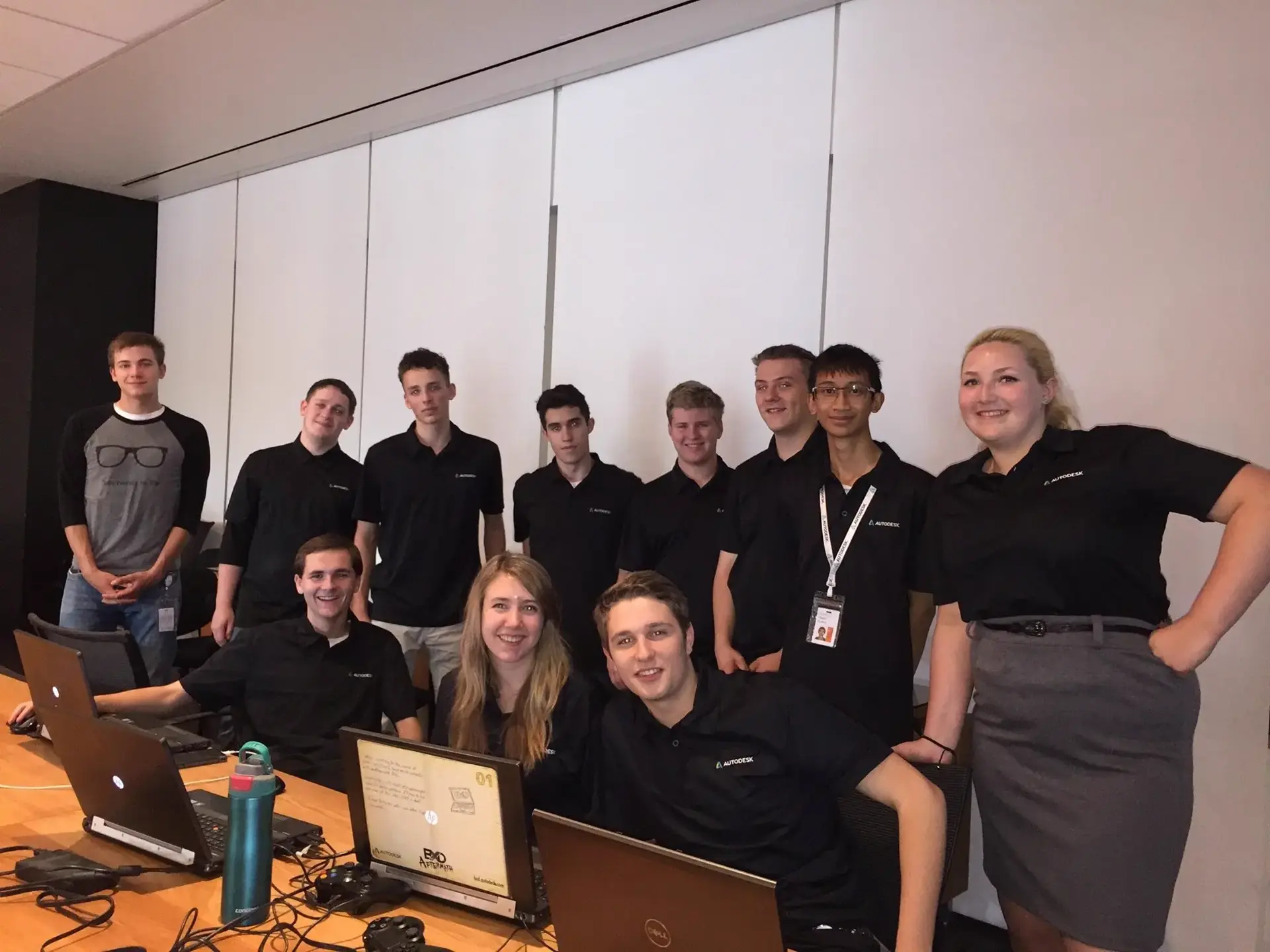 A group of 11 people looking at the camera. Most of them are wearing black Autodesk shirts. There are laptops and game controllers visible on the table in front of them.