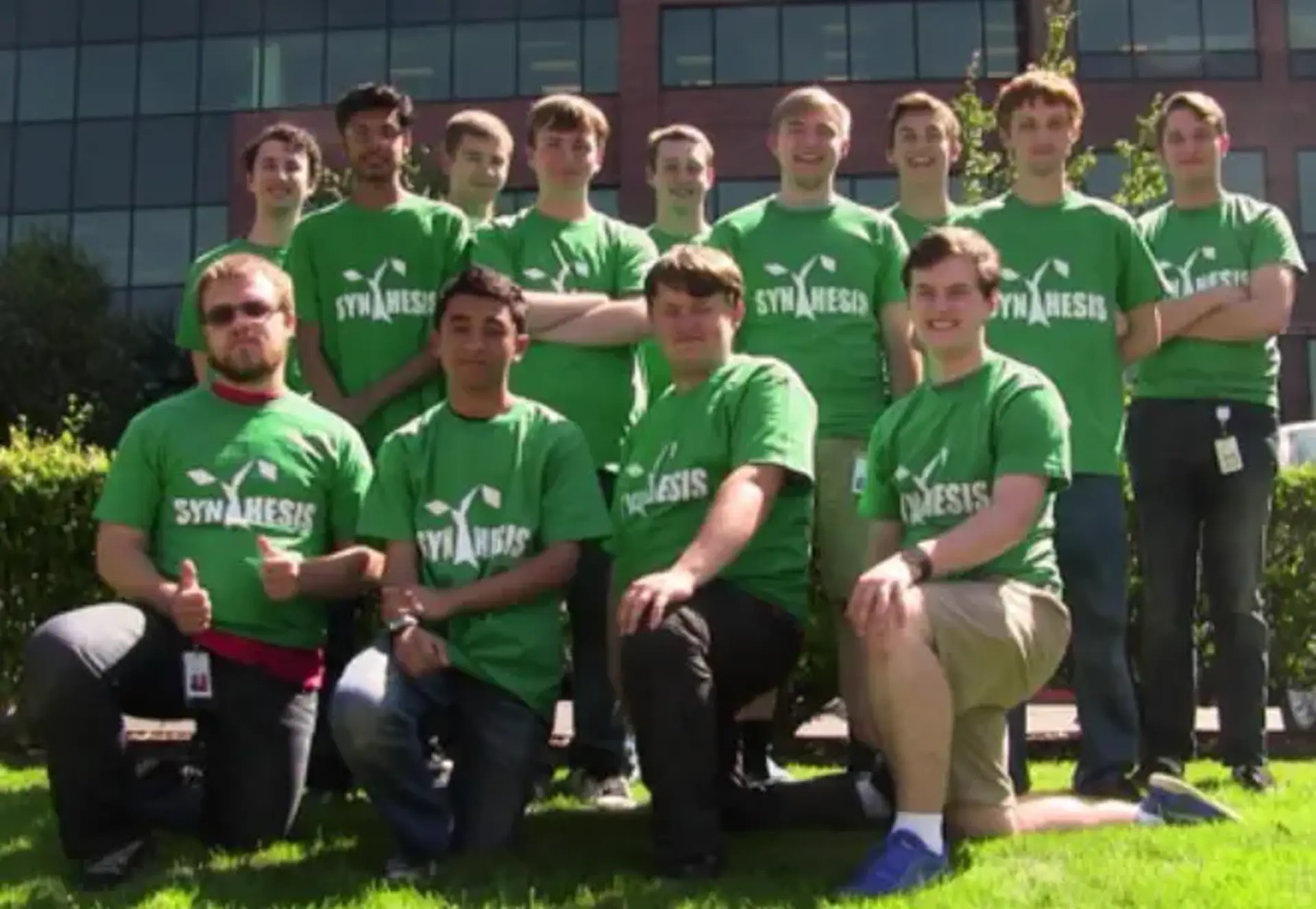 A group of 13 people on a patch of grass looking at the camera. All of them are wearing green Synthesis t-shirts.
