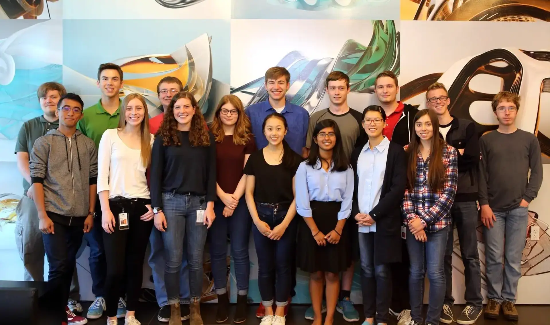 A group of 16 people standing in front of a wall all looking at the camera.