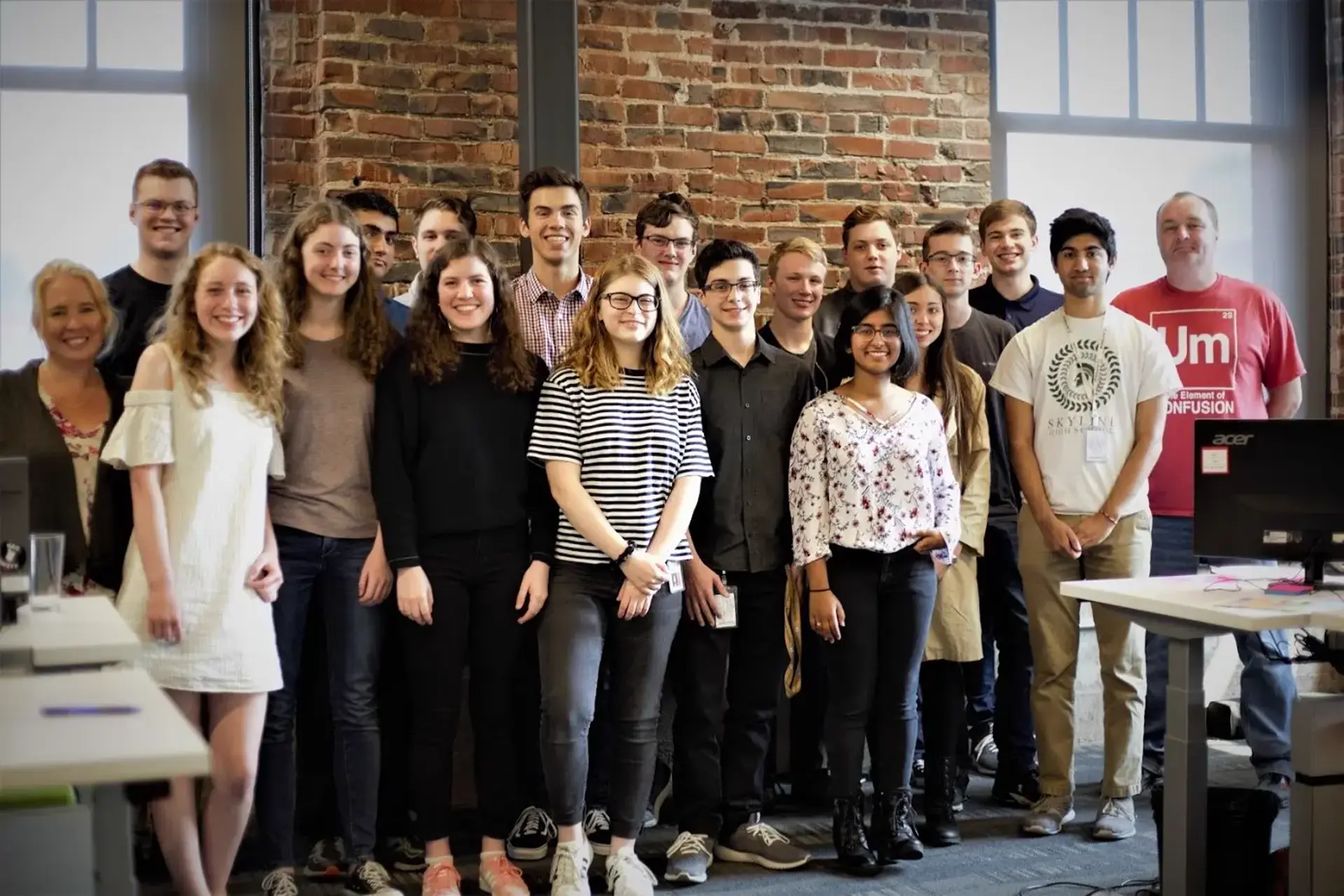 A group of 19 people looking at the camera standing in front of a wall in an office. There are desks visible on the edges of the frame
