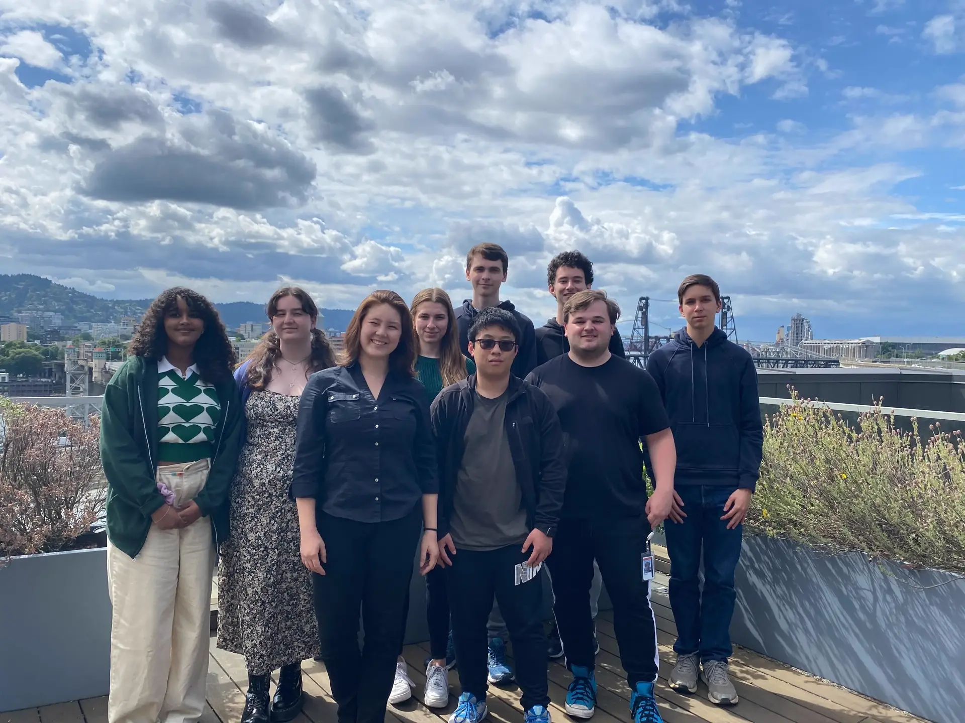 A group of 9 people looking a the camera. The photo is taking from the top of a building with downtown Portland in the background