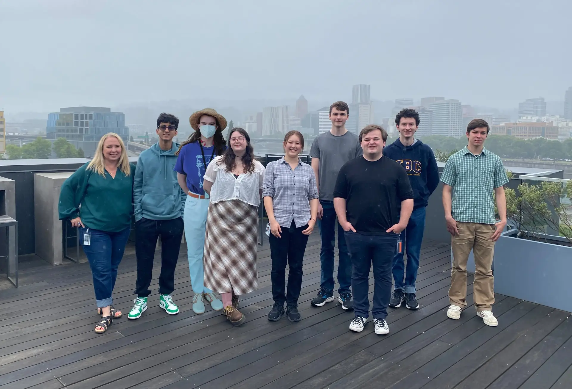 A group of 9 people looking at the camera. The photo is taken on top of a building with the downtown Portland skyline in the background