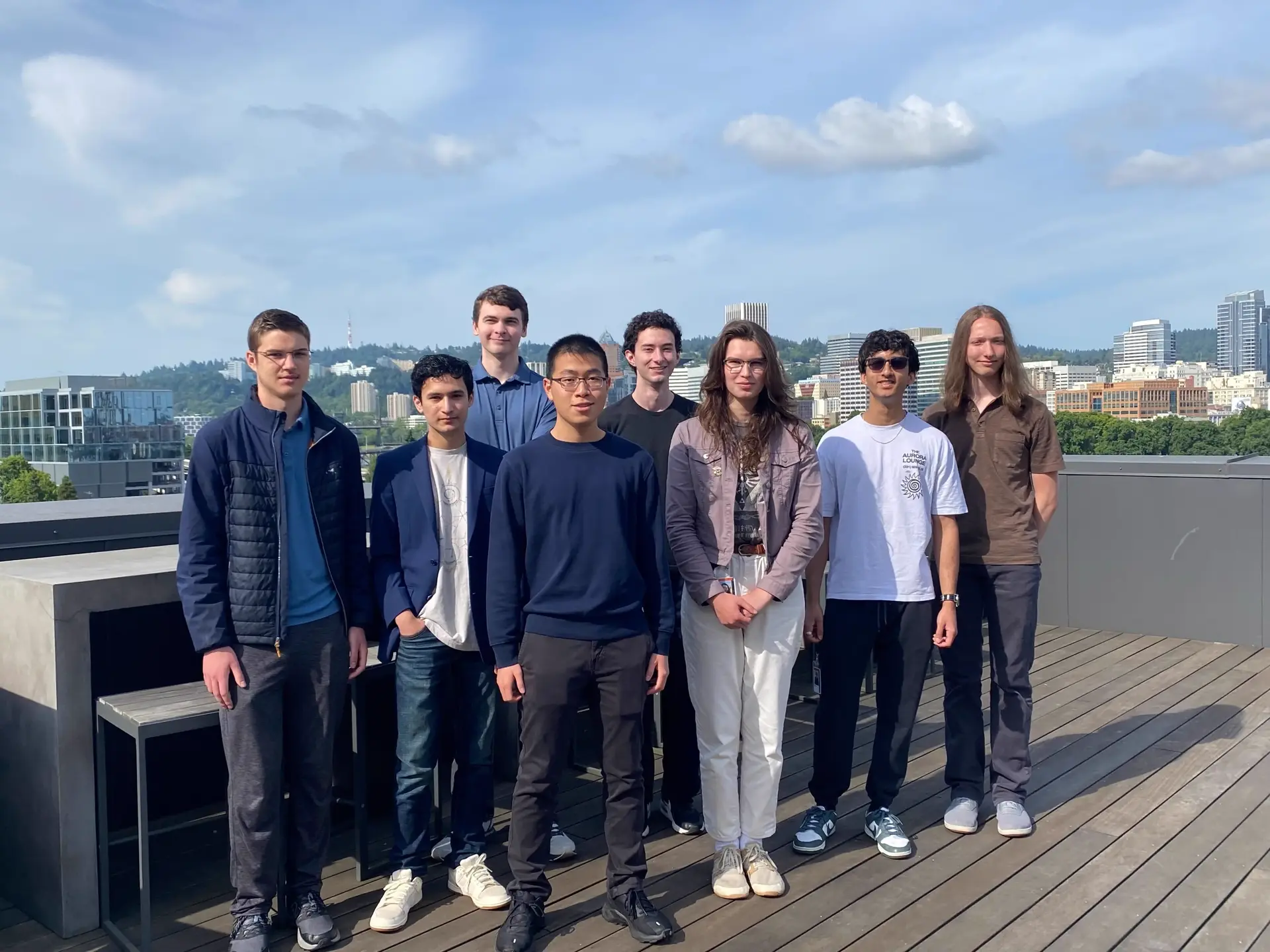 A group of 8 people looking at the camera. The photo is taken on top of a build with the downtown Portland skyline in the background.